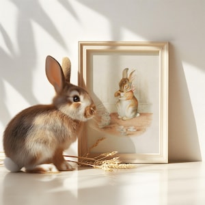 May include: A brown rabbit with white fur on its belly sits on a white surface, looking at a framed picture of a brown rabbit holding a teacup. The picture is in a light wood frame and has a white background.