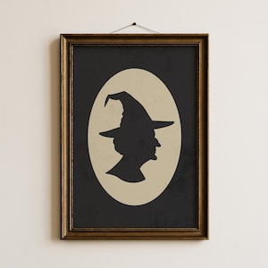 May include: Framed art print featuring a black silhouette of a witch's profile wearing a pointed hat, set against a beige oval on a dark background. The print is in a gold-colored frame, perfect for Halloween decor.