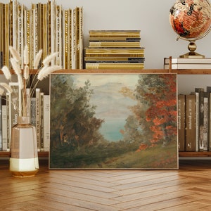 May include: A framed landscape painting with a view of a lake surrounded by trees. The painting is in a rustic style with muted colors and a soft, hazy atmosphere. The painting is hanging on a wall with bookshelves on either side.