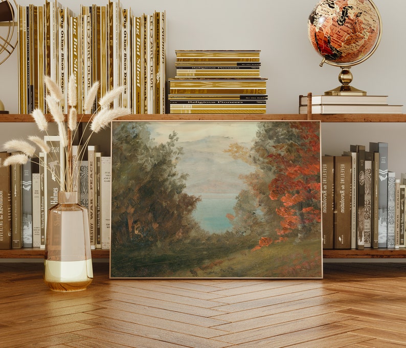 May include: A framed landscape painting with a view of a lake surrounded by trees. The painting is in a rustic style with muted colors and a soft, hazy atmosphere. The painting is hanging on a wall with bookshelves on either side.