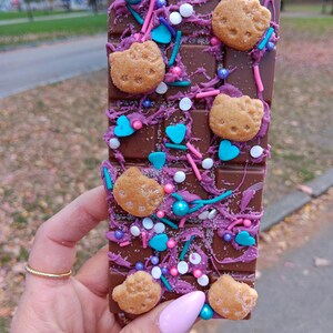 May include: A chocolate bar with a purple and pink swirl design, topped with sprinkles, hearts, and small cookie cutouts in the shape of a cat.