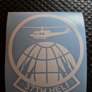 May include: A light blue sticker featuring a white helicopter above a globe design, encircled by a ring. The text "37TH HELI" is displayed at the bottom of the sticker, within a white banner.