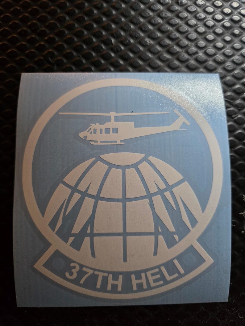 May include: A light blue sticker with a white design. It features a helicopter above a globe, encircled by a ring. The text "37TH HELI" is displayed at the bottom within a white banner.