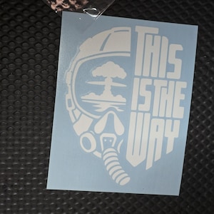 May include: A white vinyl decal featuring a stylized helmet silhouette with a mushroom cloud graphic inside the visor area. The text THIS IS THE WAY is printed in a bold, blocky font to the right of the helmet design on a light blue backing.