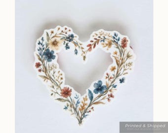 Op de afbeelding: Een hartvormige sticker met een bloemenkransontwerp van blauwe, rode en gele wilde bloemen. De tekst bovenaan luidt WILDFLOWER HEART WATERPROOF STICKER, Summer Americana, 2, 3, or 4 inches available, transparent or white.