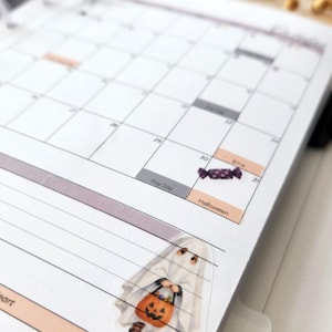 May include: A white paper planner page featuring a monthly calendar grid with printed text including Pay Day, Halloween, and Erna. A small sticker of a purple candy is placed on the 30th, and a ghost sticker holding a pumpkin bucket is below.
