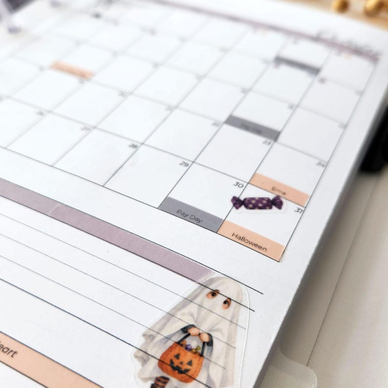 May include: A white paper planner page featuring a monthly calendar grid with printed text including Pay Day, Halloween, and Erna. A small sticker of a purple candy is placed on the 30th, and a ghost sticker holding a pumpkin bucket is below.