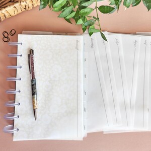 May include: A white planner with a clear plastic spiral binding and a pen with a sparkly design. The planner has tabs for each month of the year, with the month of January visible. The planner is on a light pink surface.