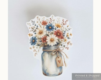 Op de afbeelding: Een waterbestendige sticker met een boeket van rode, witte en blauwe bloemen in een glazen weckpot. De pot is vastgebonden met touw en een klein beige label met een bruin hartje. Tekst op de afbeelding: GATHERED BOUQUET WATERPROOF STICKER.