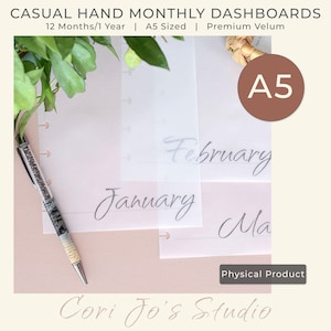 May include: A5-sized monthly dashboards with the months January, February, and March visible. The dashboards are made of premium vellum and are designed for casual hand use. A pen rests on the pink surface. The text "Cori Jo's Studio" is at the bottom.