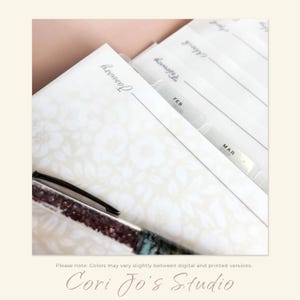 May include: A white planner with a floral pattern and clear plastic tabs labelled "January", "February", and "March". A black pen with sparkly accents is lying on top of the planner.