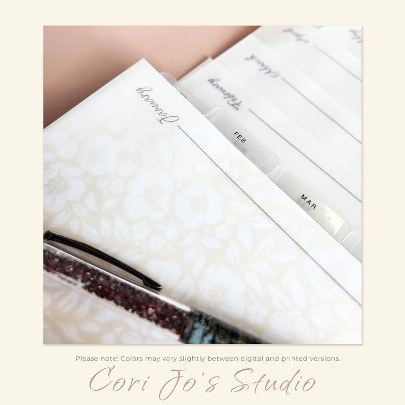 May include: A white planner with a floral pattern and clear plastic tabs labelled "January", "February", and "March". A black pen with sparkly accents is lying on top of the planner.