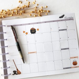 May include: A monthly planner page for October featuring a grid layout with Halloween-themed stickers including a cauldron, candy corn, a broom, and a jack-o'-lantern. A glittery pen with a dark gradient barrel rests diagonally across the planner.