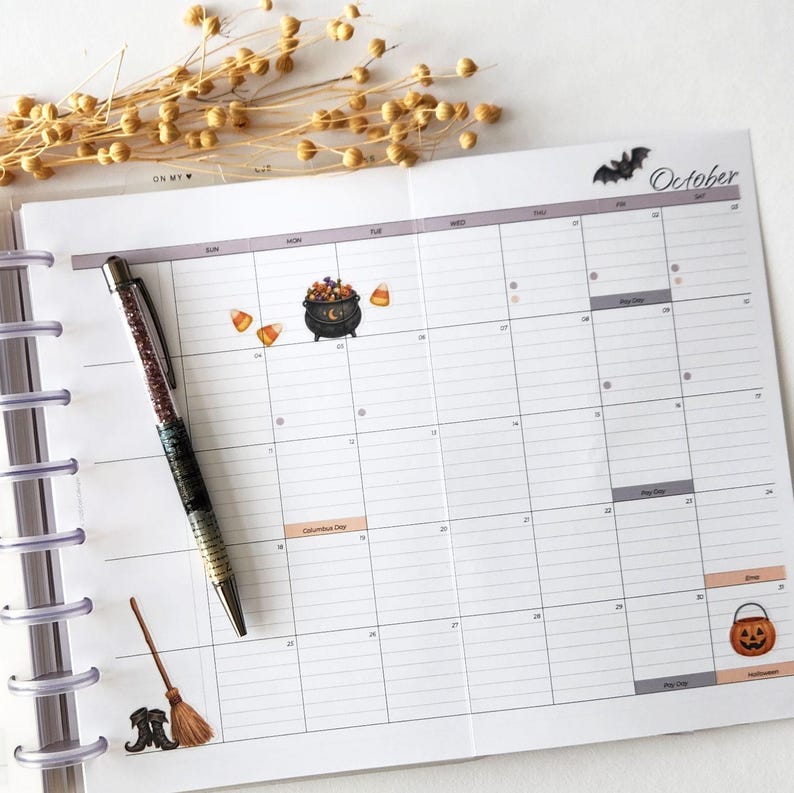 May include: A monthly planner page for October featuring a grid layout with Halloween-themed stickers including a cauldron, candy corn, a broom, and a jack-o'-lantern. A glittery pen with a dark gradient barrel rests diagonally across the planner.