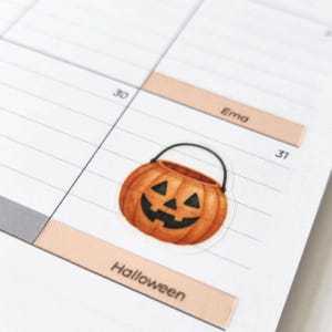 May include: A small, orange, pumpkin-shaped jack-o'-lantern sticker with a black handle and a carved face featuring triangular eyes, a triangular nose, and a jagged mouth, placed on a white paper planner page with the text Ema and Halloween.