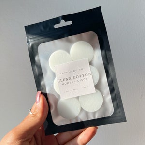 May include: A black resealable bag containing six white round scented wax melts. The bag has a label that reads "Handmade Hut Clean Cotton Hoover Discs Cruelty Free Vegan".