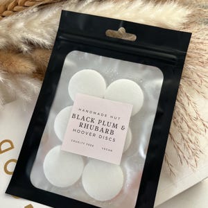 Peut inclure: Une pochette noire zipp&eacute;e avec une &eacute;tiquette blanche qui indique "Handmade Hut Black Plum & Rhubarb Hoover Discs Cruelty Free Vegan" et contient six disques ronds blancs.