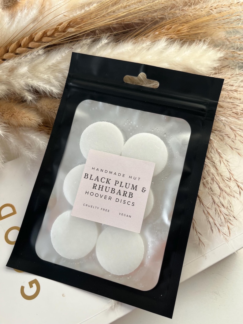 Peut inclure: Une pochette noire zipp&eacute;e avec une &eacute;tiquette blanche qui indique "Handmade Hut Black Plum & Rhubarb Hoover Discs Cruelty Free Vegan" et contient six disques ronds blancs.