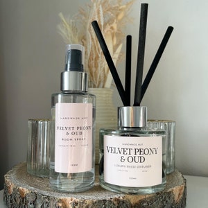 May include: Two glass bottles with silver caps and labels. The bottles contain room spray and reed diffuser. Both bottles are labelled "Velvet Peony & Oud" and are 100ml. The bottles are on a wooden surface.