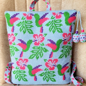 May include: A light blue crocheted backpack with a hummingbird and flower pattern. The backpack has a top handle and adjustable straps. The straps have a tassel on each end.