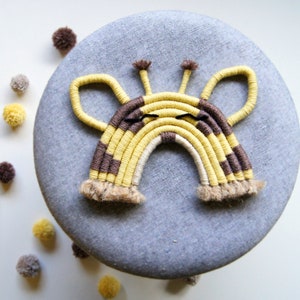 Puede incluir: Un arcoíris de macramé amarillo y marrón con una cara de jirafa, con ojos negros y una nariz marrón. El arcoíris está colgado sobre un fondo de tela gris.