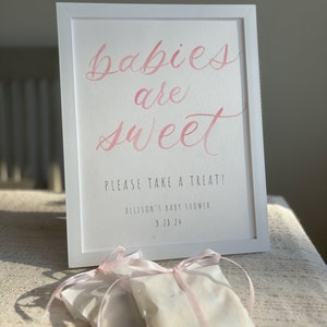 May include: A white framed sign with the text "Babies are sweet" in pink calligraphy. Below the text is "Please take a treat!" and "Allison's Baby Shower 3.23.24". There are two white bags with pink ribbons on a white surface.