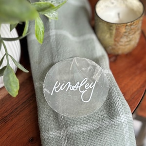 Op de afbeelding: Een ronde, transparante acryl-tafelkaart met de naam "Kingsley" geschreven in witte kalligrafie.