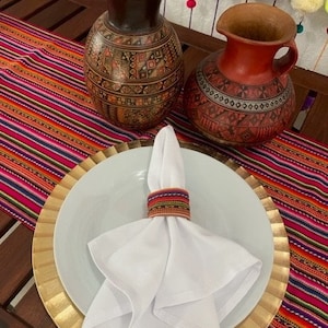 May include: A white napkin with a colorful woven napkin ring sits on a white plate with a gold rim. The plate is on top of a larger gold charger plate. The plates are on a table runner with colorful stripes. Two ceramic vases with geometric patterns are in the background.