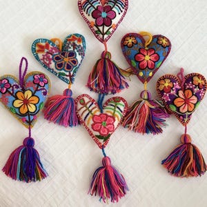 Puede incluir: Una colección de adornos bordados en forma de corazón. Cada corazón presenta un diseño floral único en colores vibrantes, con una borla a juego colgando debajo. Los adornos son de tela y están diseñados para colgar.