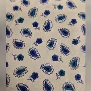 May include: Close-up of a cream-colored fabric with a repeating pattern of blue paisley teardrops and small blue floral accents. The paisley elements have a teal center and are outlined in white.