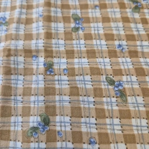 May include: A woven fabric featuring a tan and white checkered pattern overlaid with thin blue grid lines. The textile is decorated with a repeating print of small clusters of blue berries and green leaves scattered across the surface of the material.
