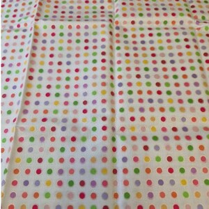 Op de afbeelding: Een witte stof met een herhalend patroon van kleine, gelijkmatig verdeelde stippen in diverse kleuren, waaronder roze, oranje, geel, groen en paars. Het textiel heeft een zichtbare geweven structuur en is in een rasterpatroon gerangschikt.
