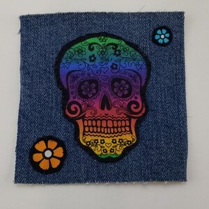 May include: A rainbow sugar skull with floral details is appliqued on a blue denim fabric background. Two small blue and orange flowers are also appliqued on the fabric.