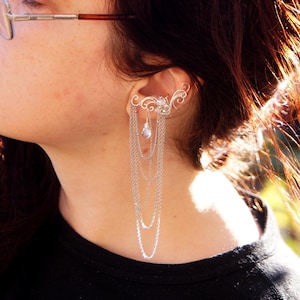 May include: A silver ear cuff with a delicate, swirling design and multiple dangling chains. The ear cuff is worn on the ear and cascades down with several strands of chain, creating a layered effect. The jewelry is a unique accessory.