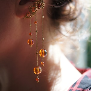 May include: Ornate gold ear cuff with dangling amber-colored beads. The ear cuff features intricate filigree designs and small red beads. The dangling beads are various sizes and hang from delicate gold chains.