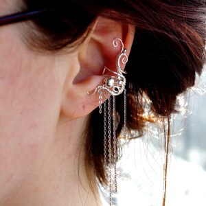 May include: Silver ear cuff with a delicate, intricate design and dangling chains. The cuff wraps around the upper ear and features a small bead.