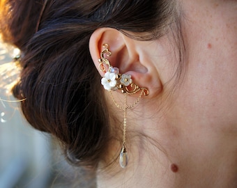 May include: A gold-toned ear cuff featuring delicate white flower accents and dangling chain with a clear teardrop-shaped charm. The ear cuff wraps around the ear with intricate filigree details, creating an elegant and whimsical look.