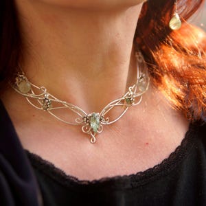 Puede incluir: Un collar de alambre plateado con una piedra preciosa verde ovalada central y pendientes a juego. El collar presenta un diseño delicado y fluido con piedras preciosas verdes más pequeñas y remolinos decorativos. Los pendientes tienen piedras preciosas verdes en forma de lágrima.