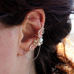 May include: A silver ear cuff with a delicate, swirling wire design. The cuff is decorated with small, round pearls and a dangling chain. The jewellery wraps around the ear and is designed to be worn without piercing.