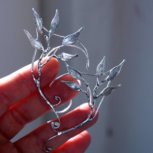 May include: A silver ear cuff with a detailed leaf and vine pattern. The ear cuff is designed to wrap around the ear, featuring multiple leaves and a curved shape. The metal has a shiny finish.