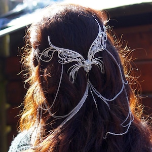 May include: A silver headpiece with a butterfly design, adorned with clear crystals and dangling chains. The headpiece is worn on dark brown hair, catching the sunlight. The jewelry piece is an elegant accessory.