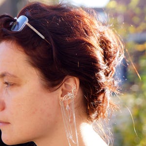 May include: A silver ear cuff with dangling chains and decorative accents. The ear cuff is worn on the ear and features a delicate design with small, dangling charms. The person in the image has auburn hair and is wearing a black top.