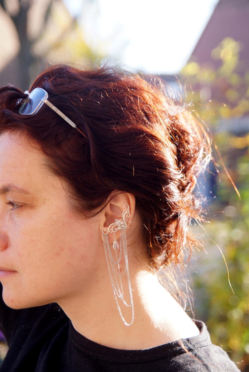 May include: A silver ear cuff with dangling chains and decorative accents. The ear cuff is worn on the ear and features a delicate design with small, dangling charms. The person in the image has auburn hair and is wearing a black top.