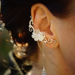 May include: A silver ear cuff featuring a floral design with white flowers and a dangling chain with a teardrop-shaped crystal. The ear cuff wraps around the ear and is worn on the ear.