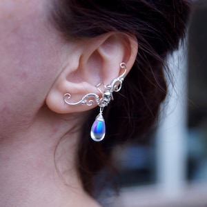 May include: Silver ear cuff featuring a detailed, ornate design. The cuff curves around the ear and has a dangling, iridescent teardrop-shaped charm. The jewelry is a unique accessory.