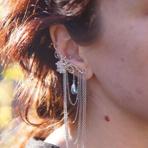 May include: A silver ear cuff featuring a floral design with dangling chains and a pearlescent bead. The ear cuff wraps around the ear and cascades down with multiple strands of delicate chains, creating a layered effect.