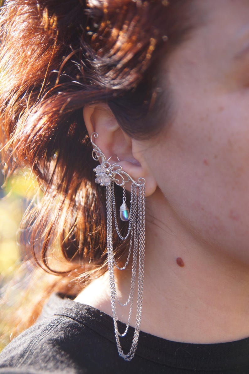 May include: A silver ear cuff featuring a floral design with dangling chains and a pearlescent bead. The ear cuff wraps around the ear and cascades down with multiple strands of delicate chains, creating a layered effect.