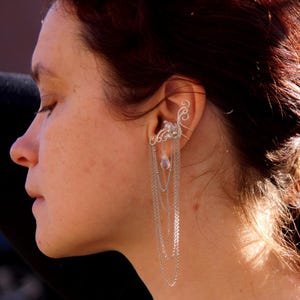 May include: Silver ear cuff with cascading chains and a small, pear-shaped crystal. The ear cuff features a delicate, ornate design that wraps around the ear. A silver hair accessory is also visible.