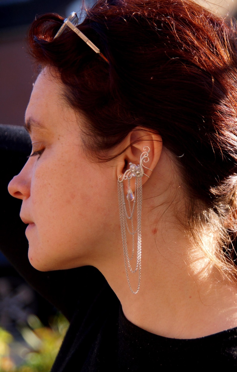 May include: Silver ear cuff with cascading chains and a small, pear-shaped crystal. The ear cuff features a delicate, ornate design that wraps around the ear. A silver hair accessory is also visible.