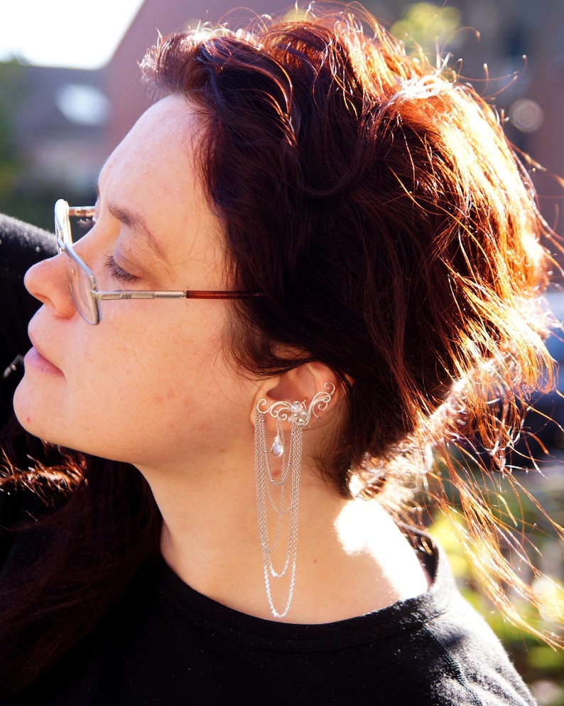 May include: A silver ear cuff with a delicate, swirling design and multiple dangling chains. The ear cuff is worn on the ear and cascades down with several strands of chain, creating a layered effect. The jewelry is a unique accessory.